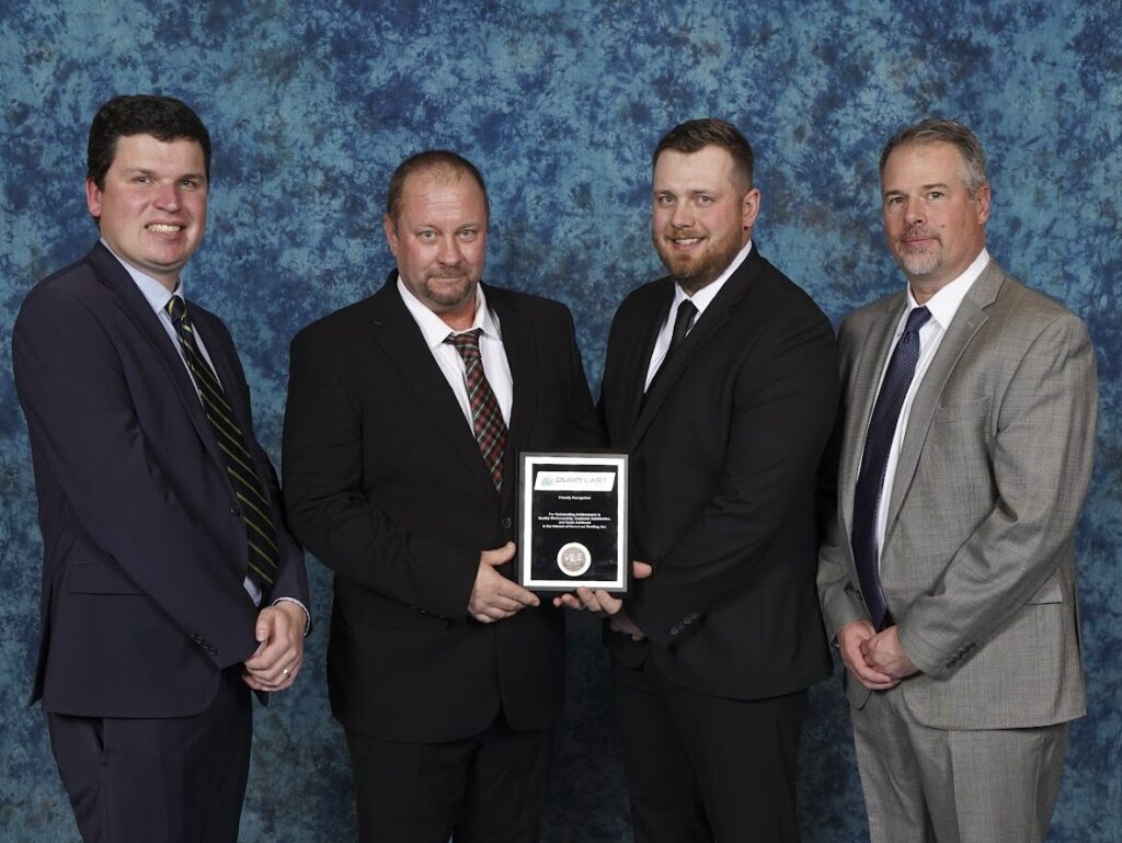 Four men stand in front of a blue background holding a plaque. They are wearing suits and smiling.